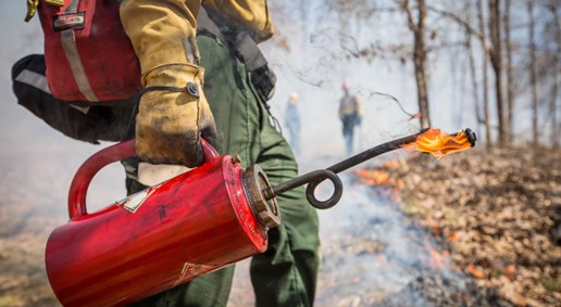 close up of a firefighter holding a lit drip torch with fire and smoke in the background
